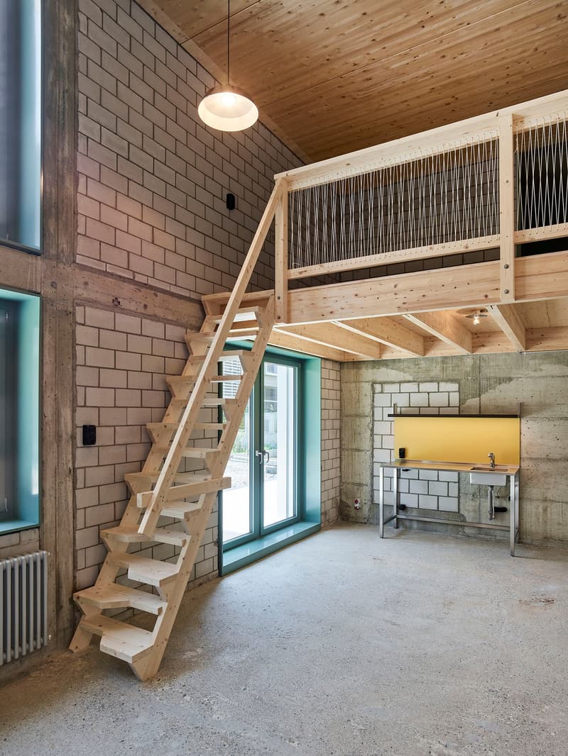 Open-concept loft-style space with exposed brick walls, wooden ceiling, and a wooden staircase leading to a mezzanine level. The space includes a kitchen area with a yellow backsplash and a window overlooking the exterior.