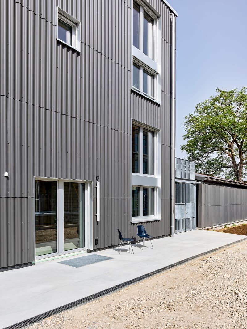 3-story building with a gray corrugated metal exterior, large windows, and a balcony on the upper floor. There are two blue chairs visible on the ground floor near the entrance.