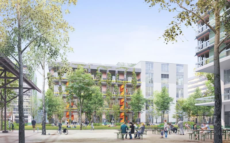 The image shows a modern residential complex with several multi-story buildings surrounded by trees and greenery. The buildings have balconies on the upper floors, and there is a central open space with people sitting at tables and benches, creating a liv