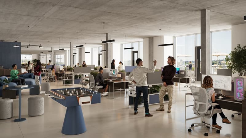 Large open-concept office space with concrete ceiling, modern lighting fixtures, and floor-to-ceiling windows providing a view. The space features a mix of workstations, lounge areas, and a foosball table. The office appears to have a casual, collaborativ