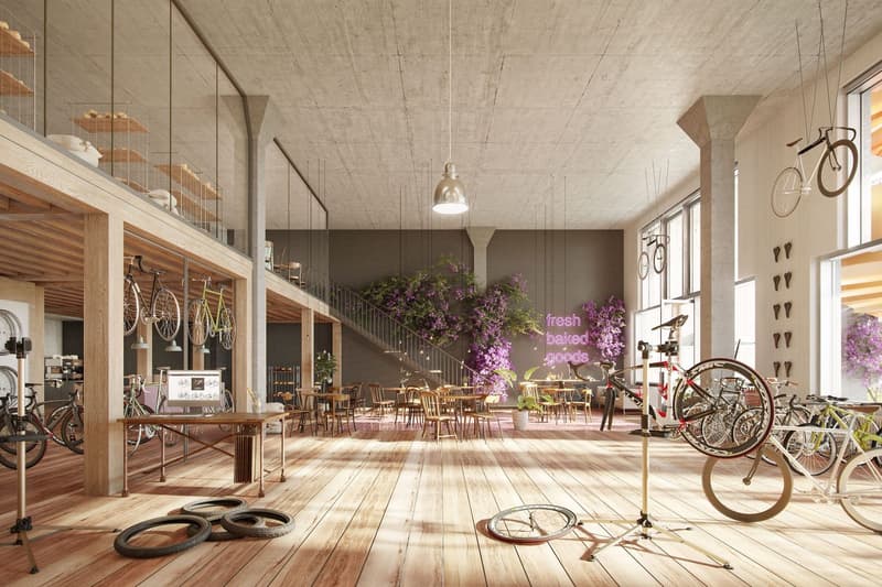 Large open space with concrete ceiling, wooden floors, and shelves displaying various bicycle parts and accessories. The space features a seating area with tables and chairs, as well as a counter area with a 'fresh baked goods' sign. Several bicycles are