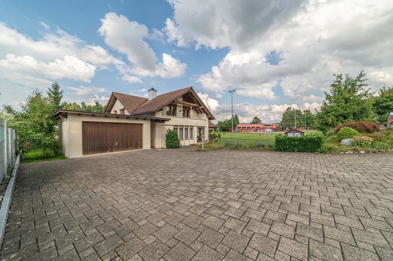 two-story house, brown roof, wooden garage, paved driveway, green lawn, trees, bushes, street light