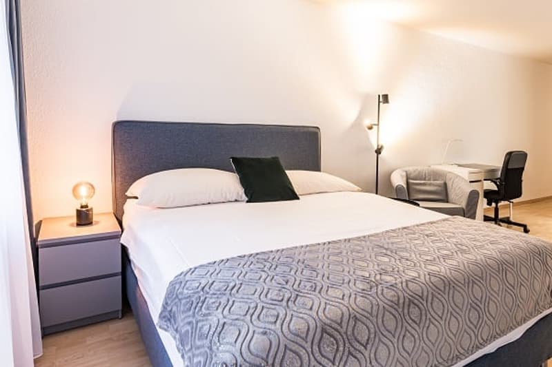 Spacious bedroom with a large bed, bedside tables, and a chair. The room has large windows that provide natural light and a cozy atmosphere.