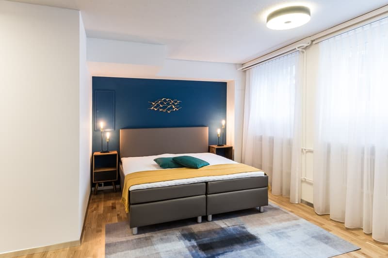 Spacious bedroom with a large bed, dark blue accent wall with decorative birds, bedside tables, and a large window with curtains.