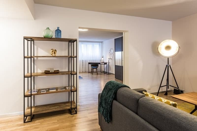 Spacious living room with hardwood floors, white walls, and a large window. The room features a wooden shelving unit, a gray sofa, and a floor lamp. The doorway in the background suggests an open floor plan leading to other rooms.