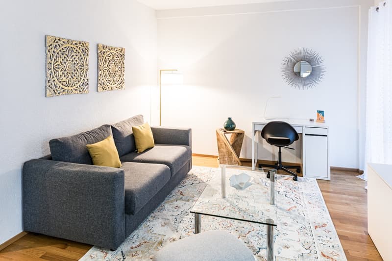 Modern room with a sofa, carpet, glass table, desk, chair, lamp, and wall art.