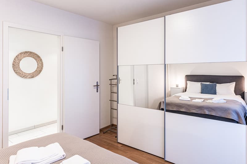 A small bedroom with wooden floor, a bed with pillows, a closet with a mirror, a nightstand with a lamp, and a small round decorative wall mirror.