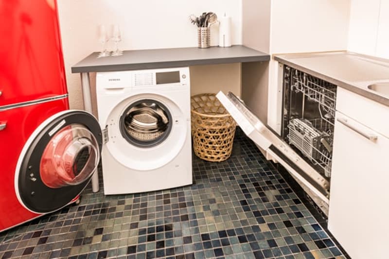 Red refrigerator, white washing machine, silver dishwasher, wicker basket, tiled floor, white cabinets, silver faucet