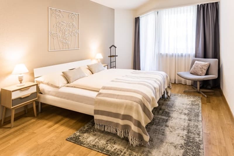 a well-decorated bedroom with a large bed, a rug, a nightstand with a lamp, a picture frame on the wall, a chair, and a small table with a lamp