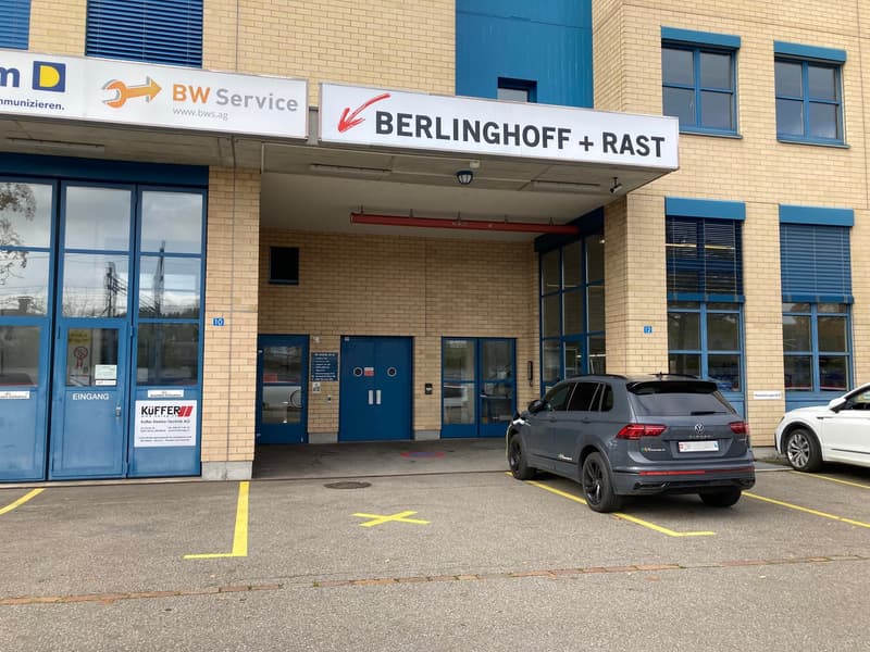 2-story commercial building with a BW Service sign and a Berlinghoff + Rast sign. The building has a blue entrance door and large windows. There is a Volkswagen SUV parked in the parking lot in front of the building.