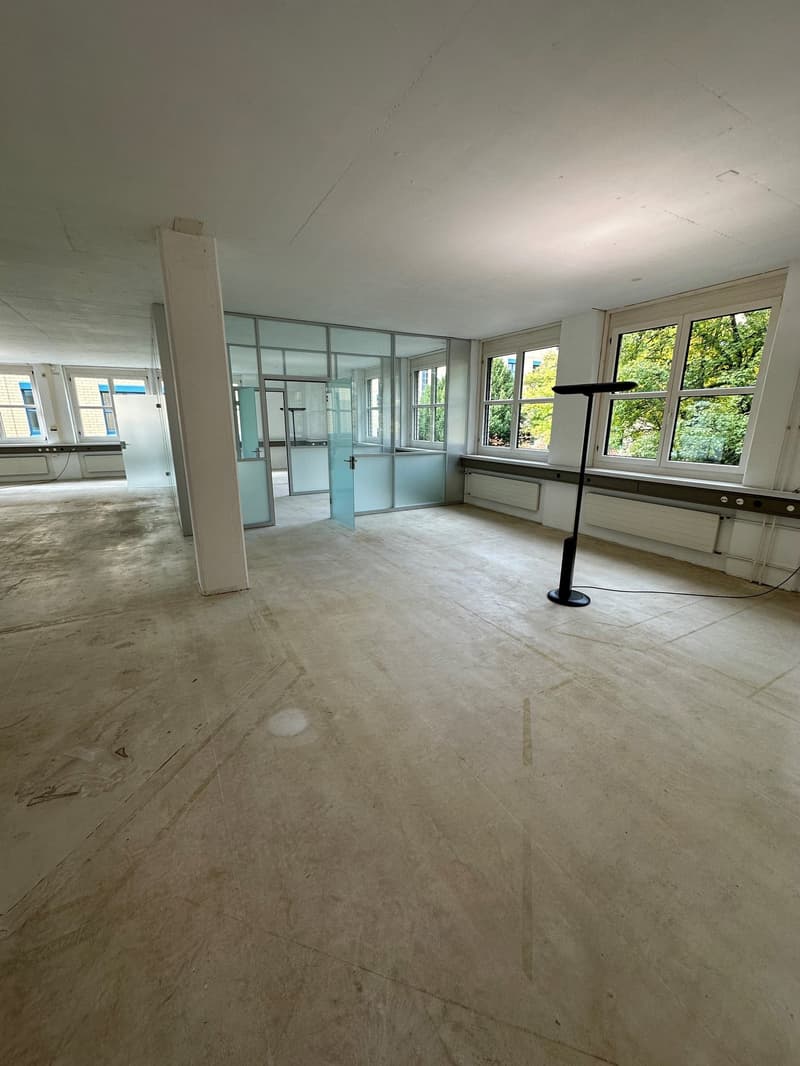 The image shows a large, open office space with concrete floors, glass walls, and large windows overlooking greenery outside. The space appears to be in the process of renovation or refurbishment, with minimal furnishings and equipment visible.