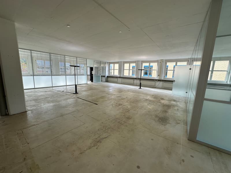 Large open-plan office space with concrete flooring, high ceilings, and large windows providing natural light. The space appears to be in a state of renovation or construction, with minimal furnishings and equipment present.