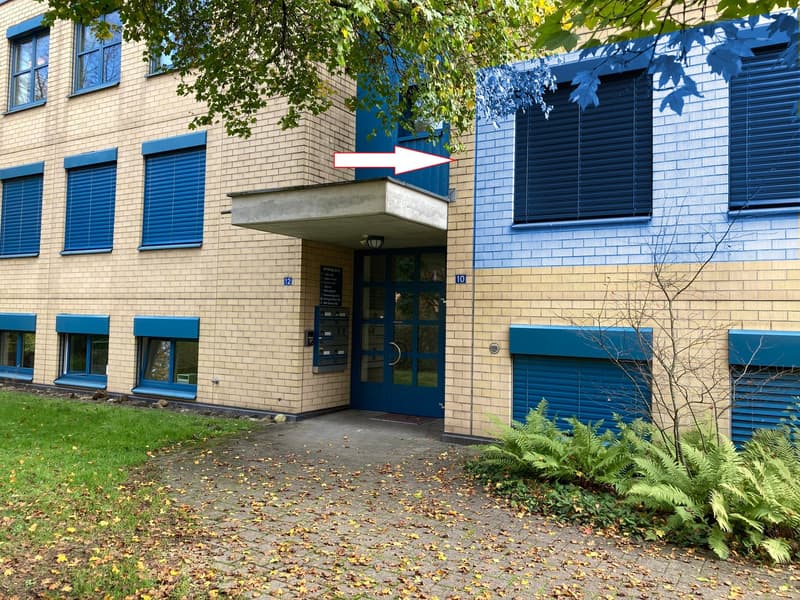 2-story brick building with blue windows and shutters, entrance with a concrete overhang, surrounded by trees and plants