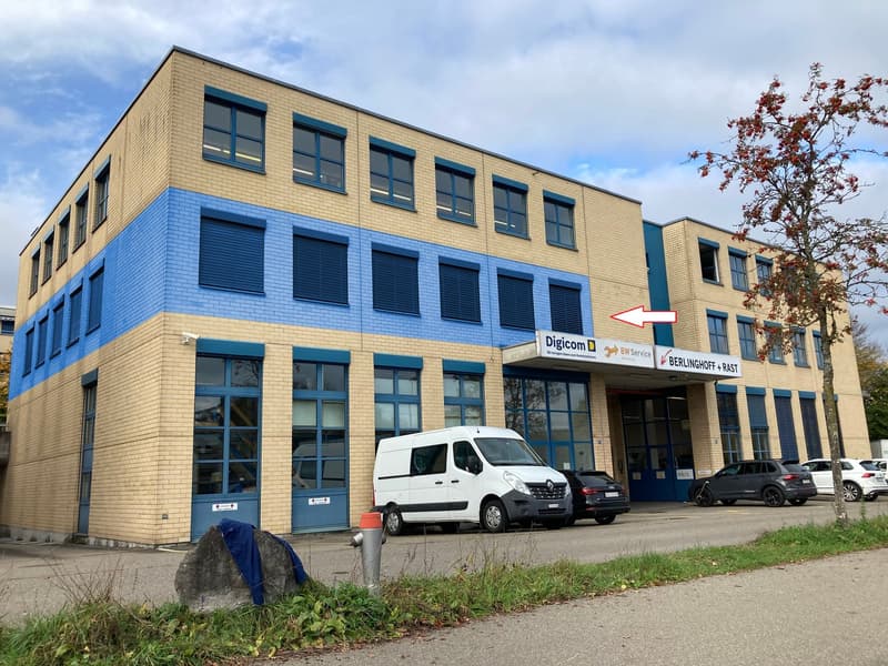 3-story commercial building with yellow and blue facade, large windows, Digicom and Berlinghoff & Rast signage, parking spaces in front of the building