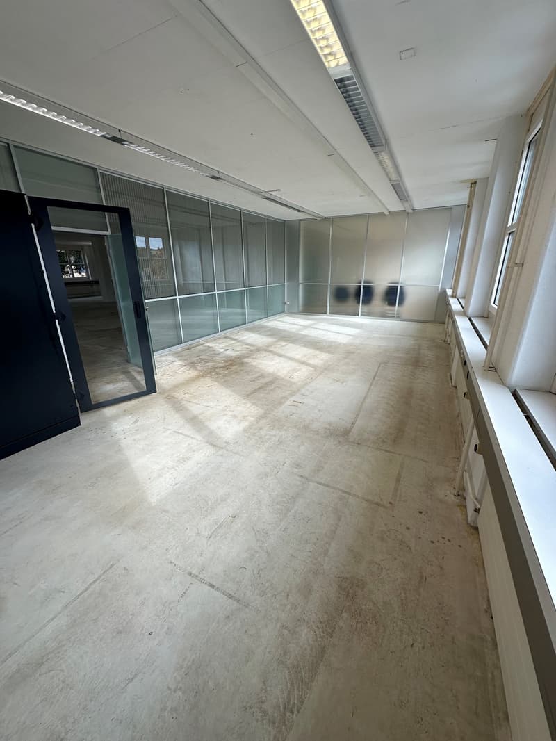 Large open office space with concrete flooring, glass partition walls, and overhead lighting. The space appears to be in a state of construction or renovation.