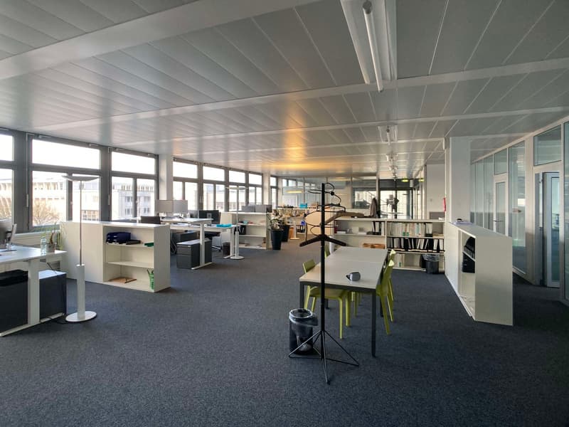 Open office space with cubicles, desks, computers, and shelving units. Large windows provide natural light. The space has a modern, minimalist design with a gray carpet floor and a white ceiling with exposed beams.