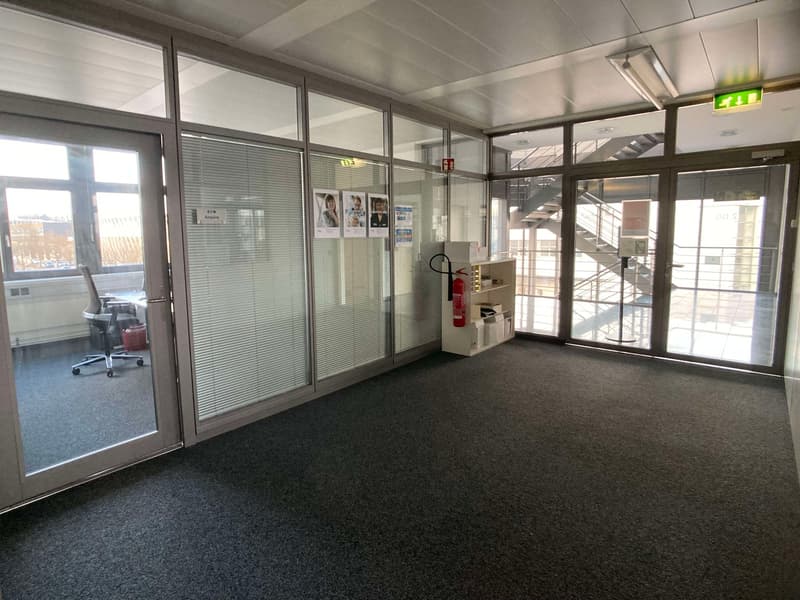 The image shows an office space with glass partitions, a carpeted floor, and a fire extinguisher cabinet. The space has a professional and functional appearance, with a desk and chair visible through one of the glass partitions.