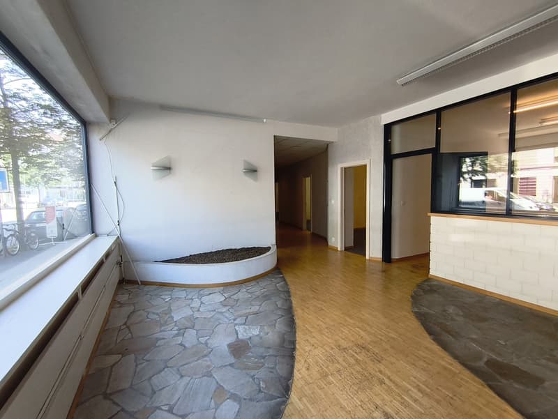 Large open-plan room with hardwood floors, stone accent wall, and large windows providing natural light. The room appears to be a living or dining area with a raised platform or seating area in the corner.