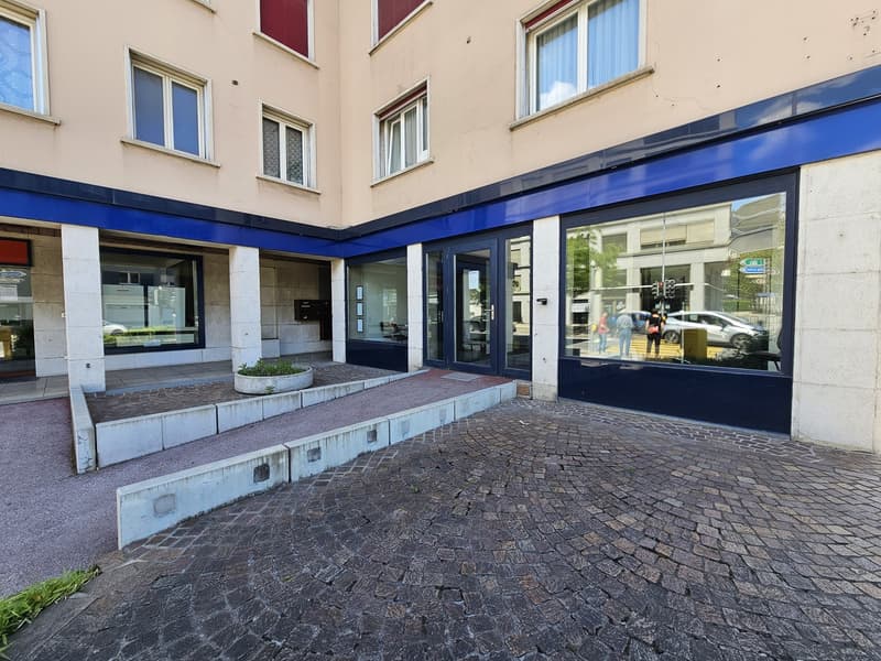 A multi-story commercial or residential building with a blue-framed glass entrance and windows. The building has a beige exterior and a cobblestone walkway leading to the entrance. There are people visible through the glass windows.