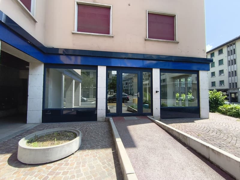 The image shows a commercial building with a blue awning and large glass windows. The building has a beige exterior and there is a raised concrete platform with a planter in front of the entrance. The surrounding area appears to be a residential neighborh