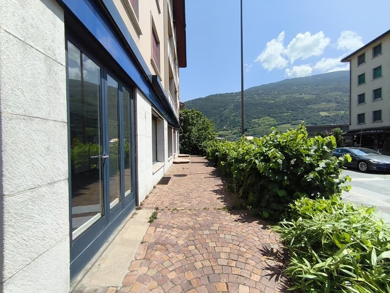 Multi-story commercial or residential building with large windows, brick paved walkway, and lush greenery surrounding the building.