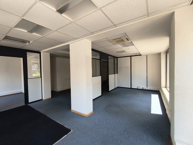 The image shows an empty office space with a suspended ceiling, white walls, and a gray carpeted floor. The room has several glass-paneled partitions dividing the space into different sections. There is an air conditioning unit visible on the ceiling.