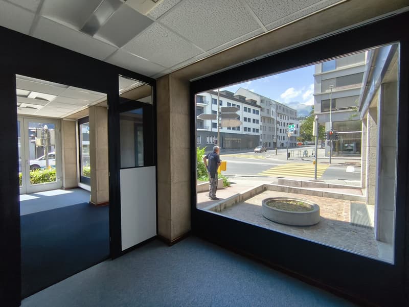 The image shows an interior office space with large windows overlooking a city street. The space has a tiled ceiling, gray carpeted floor, and black walls. There is a person visible through the window, standing on the sidewalk outside the building.