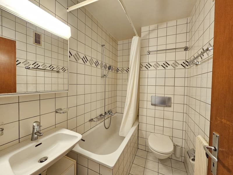 Tiled bathroom with a bathtub, sink, toilet, and shower curtain. The walls are covered in white tiles with a decorative pattern.