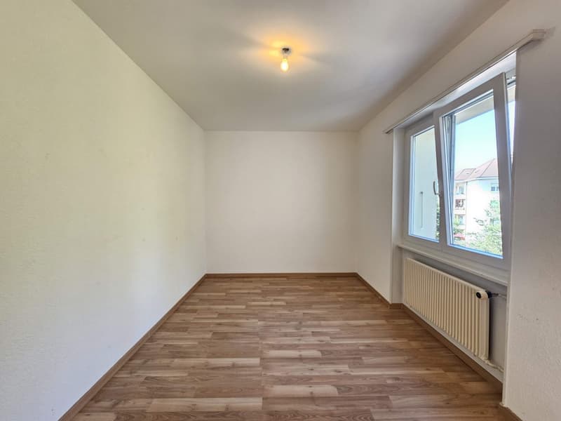 The image shows a spacious bedroom with white walls and a hardwood floor. The room has a large window that provides natural light and a view of the surrounding buildings. There is a single light fixture on the ceiling.