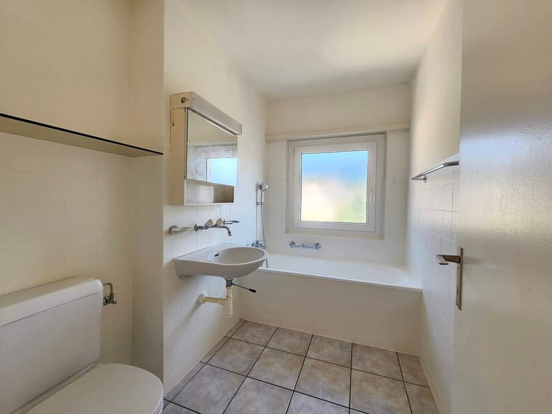 The image shows a bathroom with a white sink, a bathtub, and a window. The walls are tiled, and the floor is covered with square tiles. The bathroom appears to be clean and well-maintained.