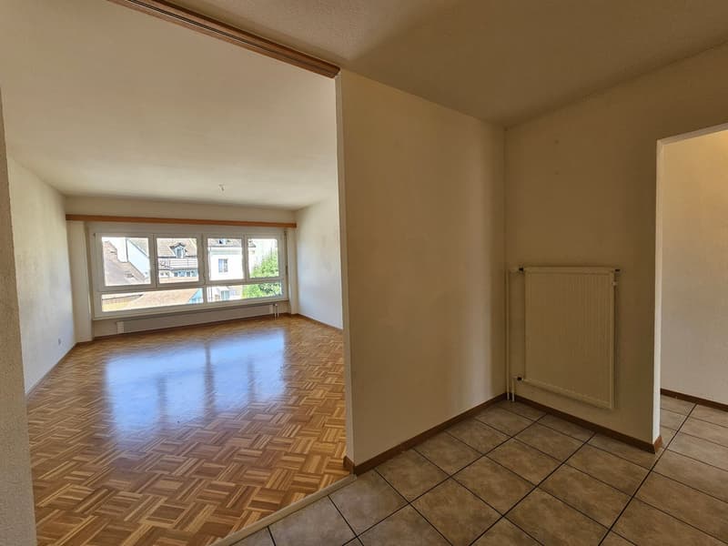 Spacious living room with hardwood flooring, large windows, and a radiator. The room has beige walls and appears to be part of a larger apartment or home.