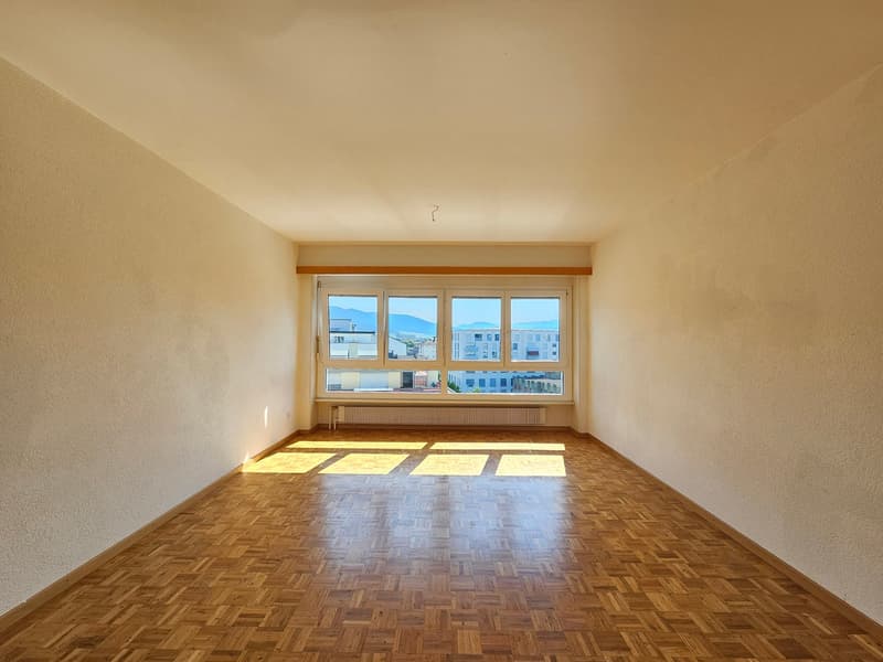Spacious living room with large windows, hardwood parquet flooring, and a wooden trim along the ceiling.