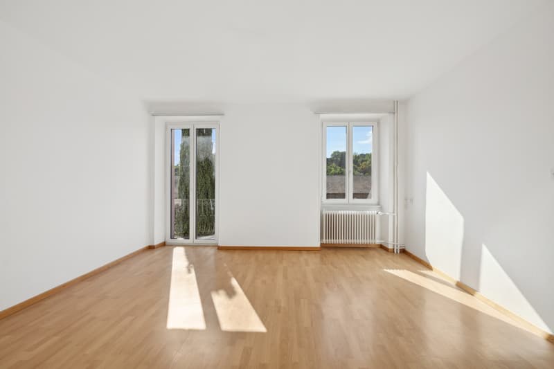 Spacious room with large windows, hardwood flooring, and a radiator. The room appears to be empty, with plenty of natural light streaming in.