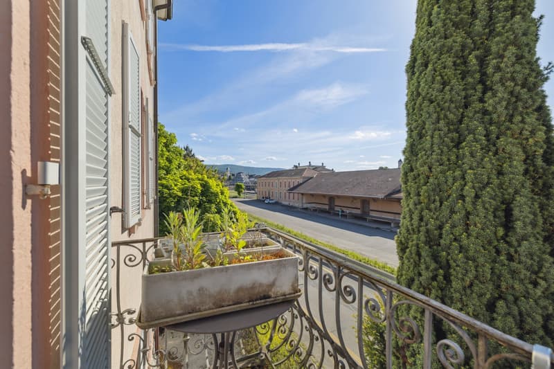 The image shows the exterior of a building with a balcony or terrace. The building has shuttered windows and a wrought-iron railing on the balcony. There are tall evergreen trees lining the street in the background, and the sky is blue with some clouds.
