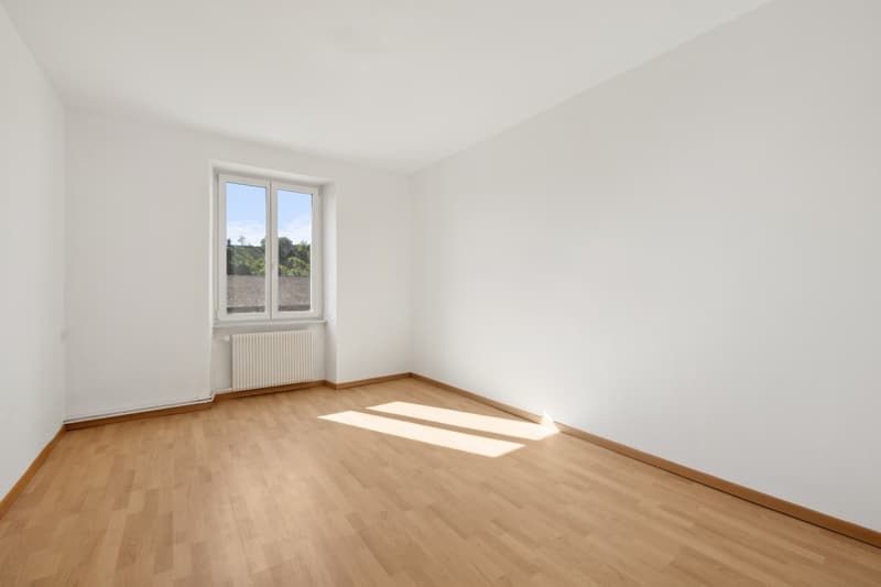 Spacious bedroom with white walls, large windows, and hardwood flooring. The room appears bright and airy, with natural light streaming in through the windows.