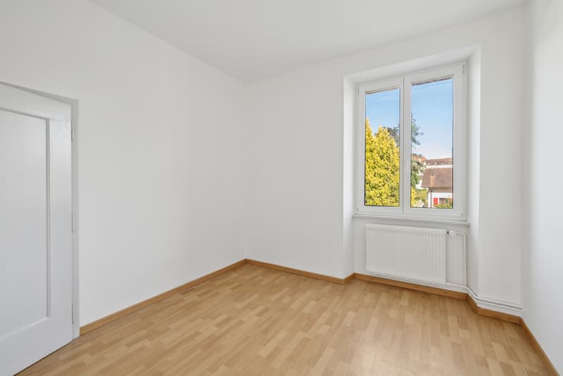 Spacious bedroom with white walls, hardwood flooring, and large windows overlooking a view of trees and buildings outside.