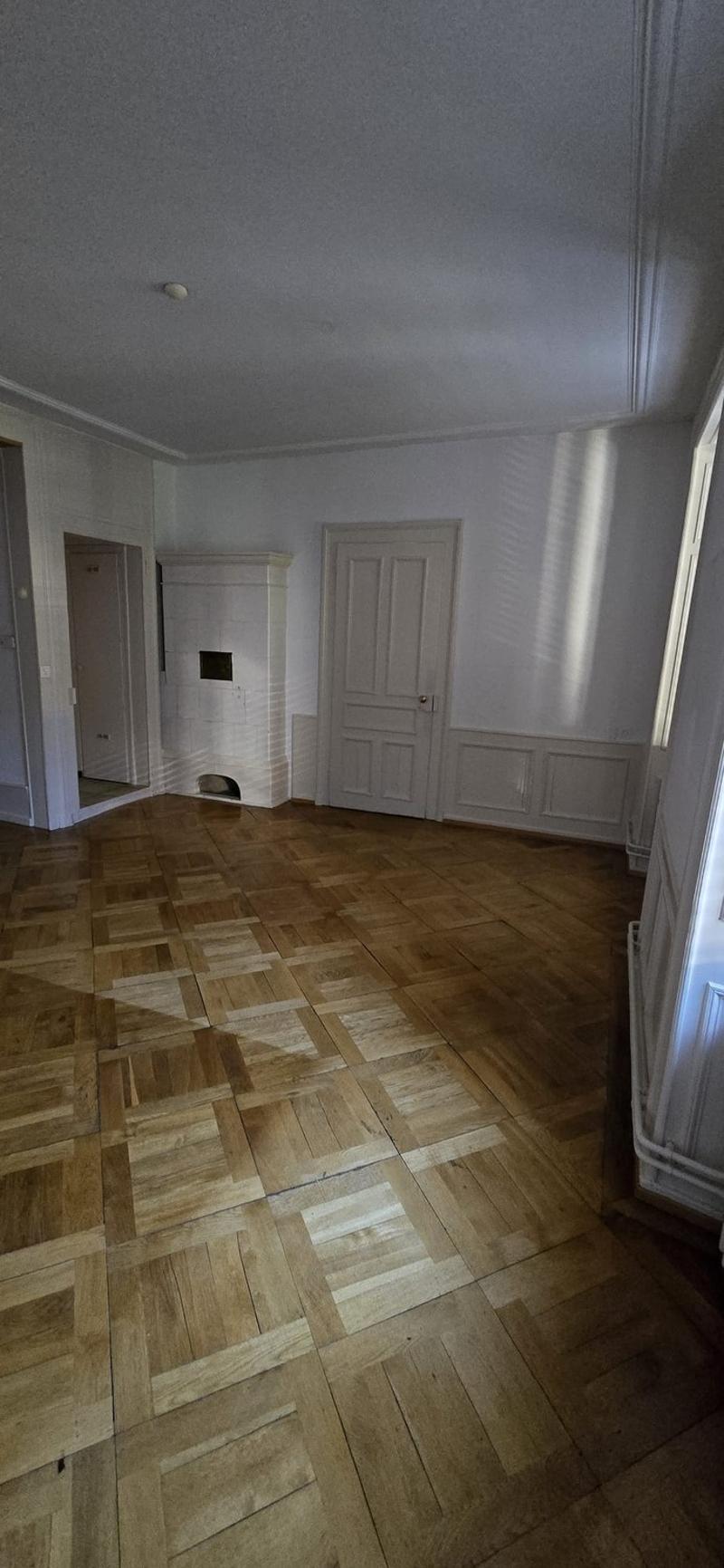 The image shows a room with white walls, a wooden parquet floor, and a white door. The room appears to be empty and has a minimalist design.