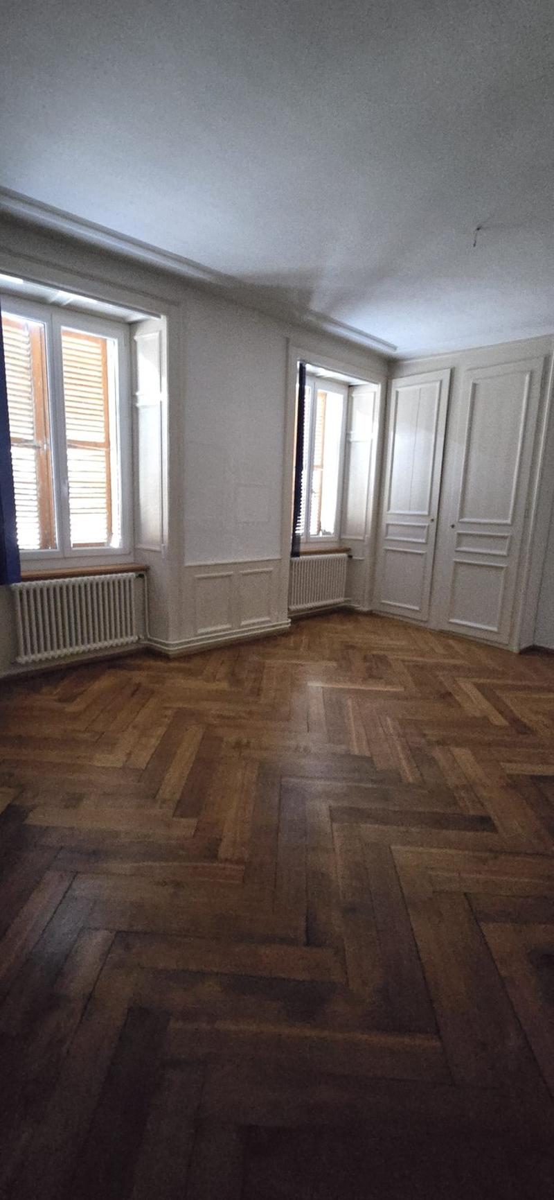 This image shows a room with white walls, large windows, and a hardwood floor in a herringbone pattern. The room appears to have built-in cabinets or closets along one wall. There are radiators visible, indicating this is likely a residential space.