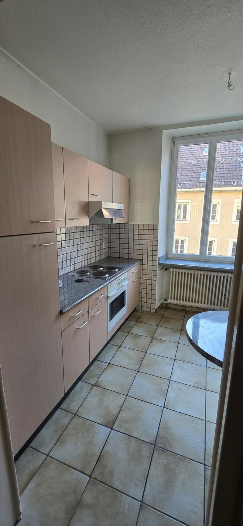Compact kitchen with light-colored cabinets, tiled backsplash, and a dishwasher. The kitchen has a window overlooking the exterior buildings.