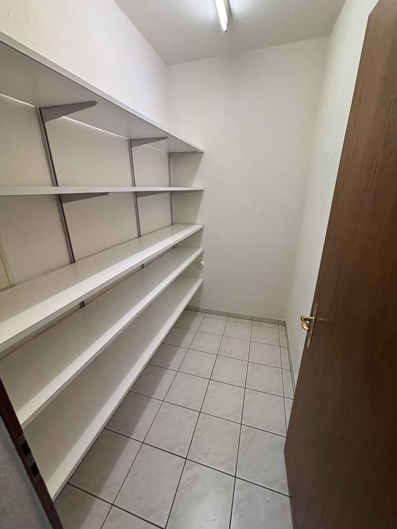 The image shows a small closet or storage room with white shelves and a tiled floor. The room appears to be well-lit and has a wooden door.