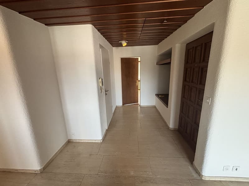 The image shows a long hallway with a wooden ceiling and tile flooring. The walls are painted a light color, and there are several doors leading to other rooms. The hallway appears to be part of a larger residential or commercial space.