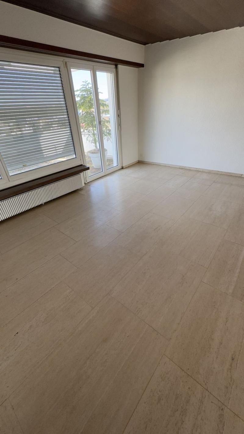 This image shows an empty room with a large window and tile flooring. The walls are painted a light color and there are wooden beams on the ceiling. The room appears to be a living room or bedroom based on the size and layout.