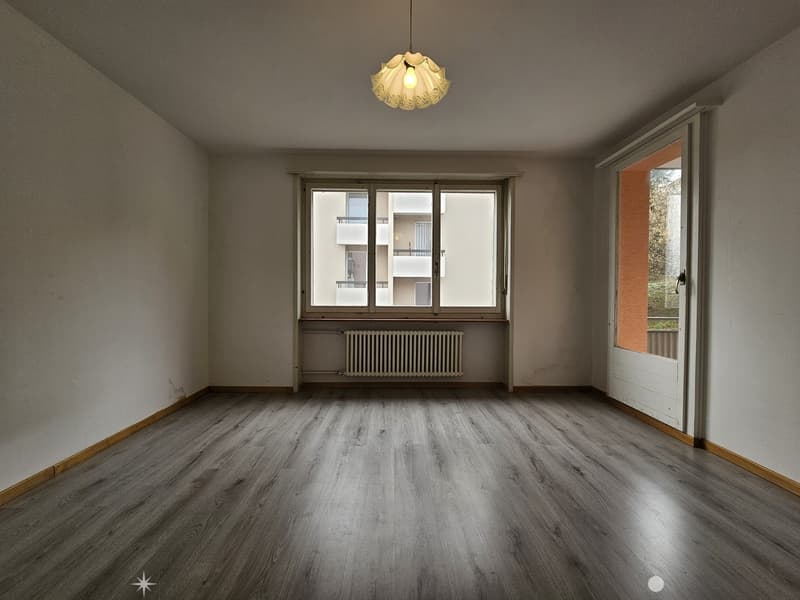 Spacious living room with hardwood flooring, large windows, and a decorative pendant light fixture. The room appears to be in a residential apartment or condominium setting.