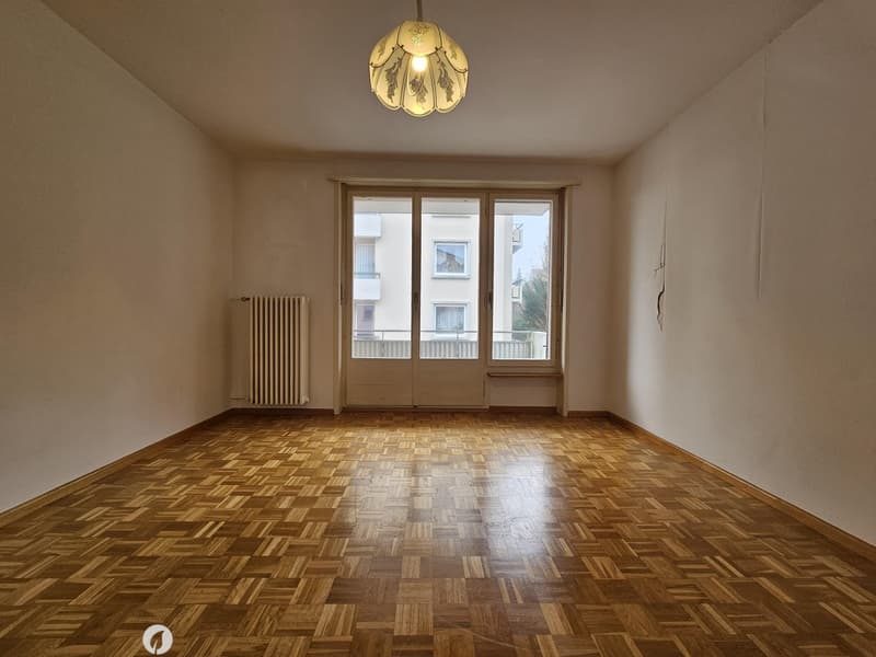 This image shows a spacious room with hardwood parquet flooring, a large window with a radiator underneath, and a decorative light fixture on the ceiling. The walls are painted a neutral color, creating a warm and inviting atmosphere.
