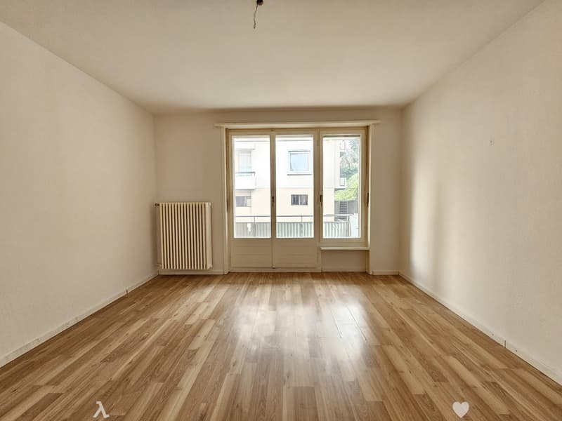 Spacious living room with hardwood floors, large windows, and a radiator. The room appears to be empty, ready for furnishing and decorating.