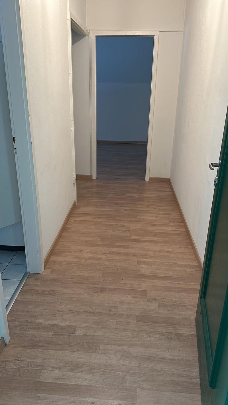 The image shows a hallway with white walls and a wooden floor. The hallway leads to a room with a blue wall visible through the open doorway.