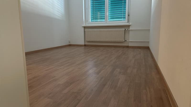 Empty room with hardwood flooring, white walls, and a radiator under the window with closed blinds.