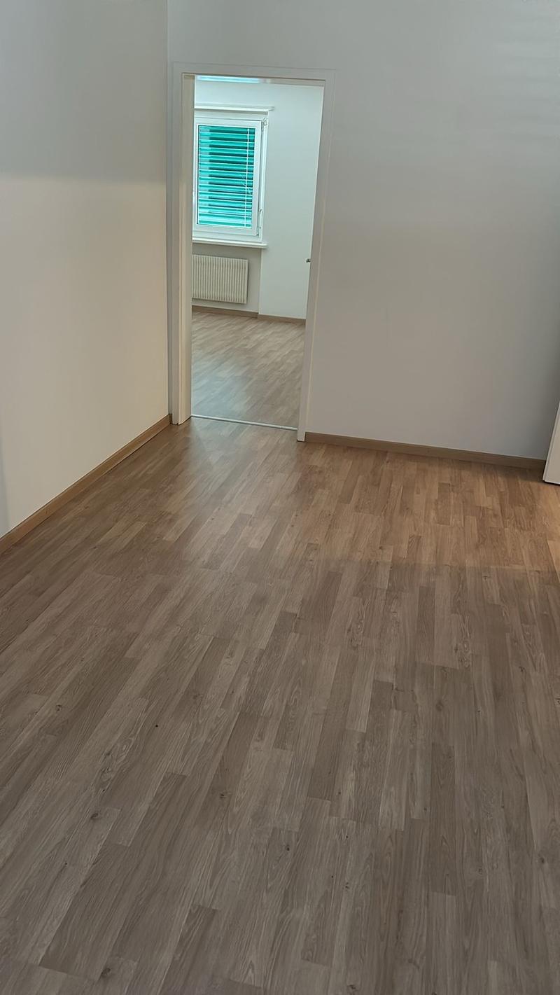 Empty room with light gray walls and light wood laminate flooring. A window with green blinds is visible in the adjacent room.