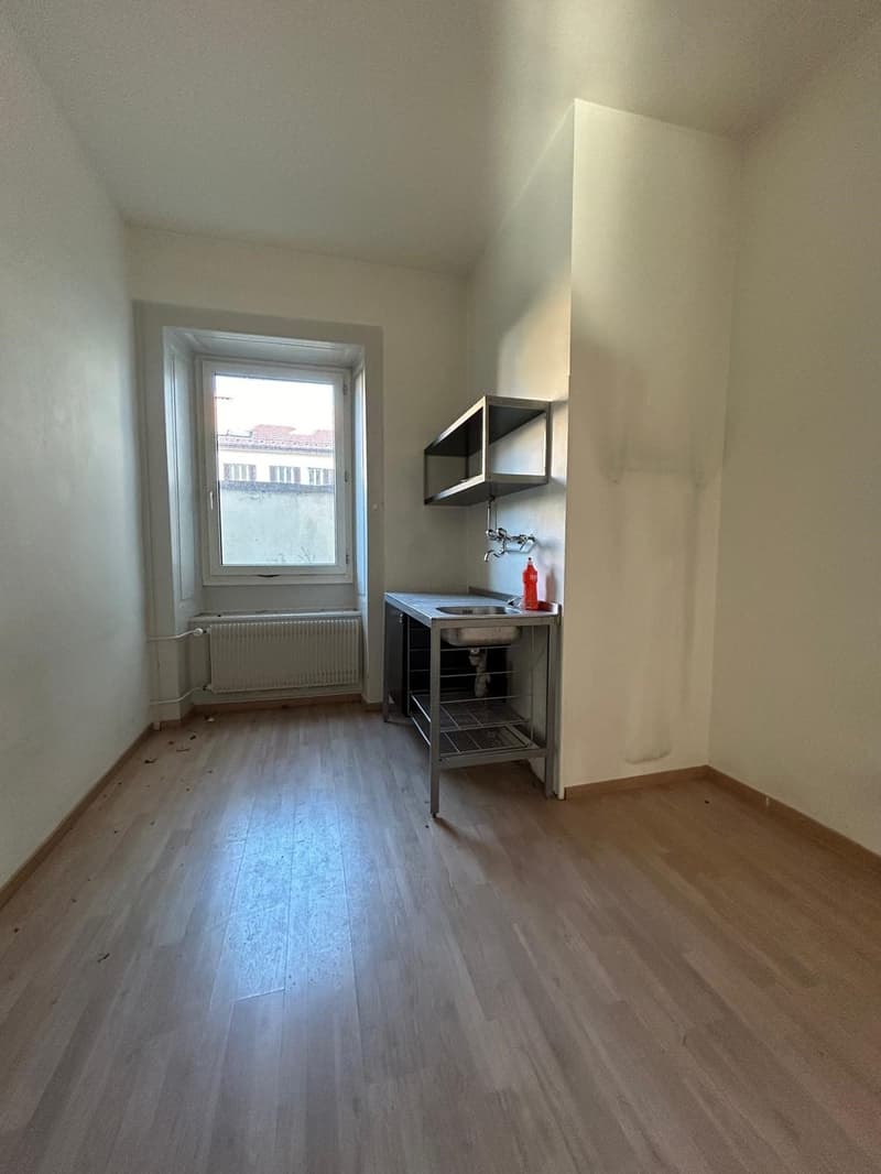 The image shows a small room with a window, hardwood floors, and a metal shelving unit with a sink. The room appears to be a kitchenette or small kitchen area within a larger living space.