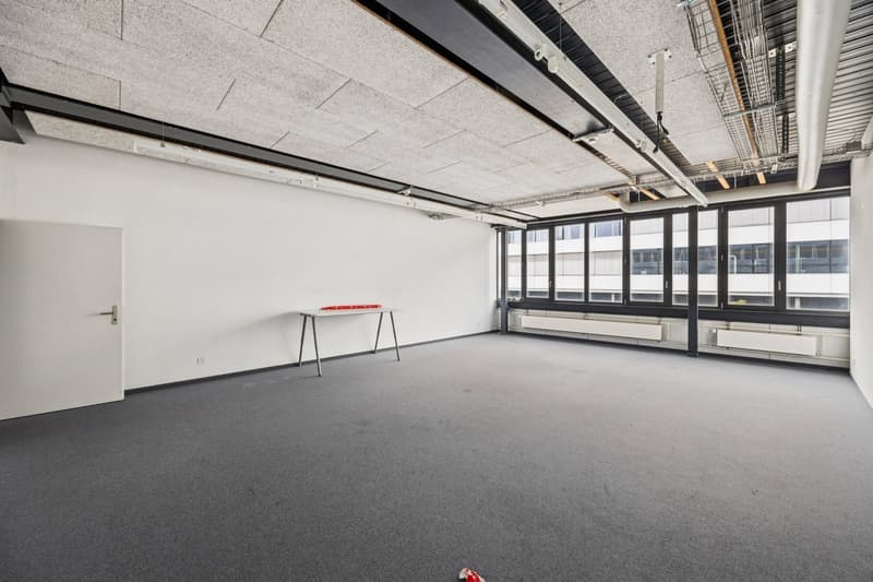 Large open office space with concrete ceiling, exposed ductwork, and gray carpeted floor. The room has large windows providing natural light and a small table near the entrance.